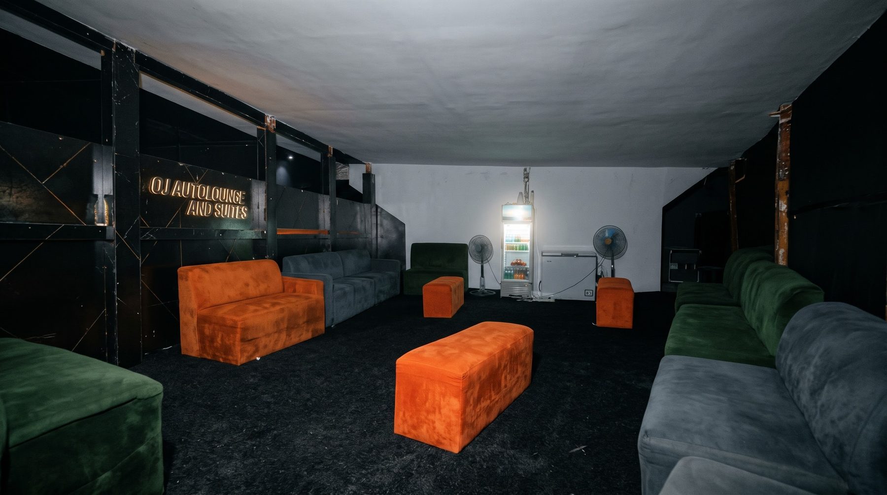 The lounge with orange, green and grey seating, the OJ Autolounge and Suites wall sign and a drinks fridge